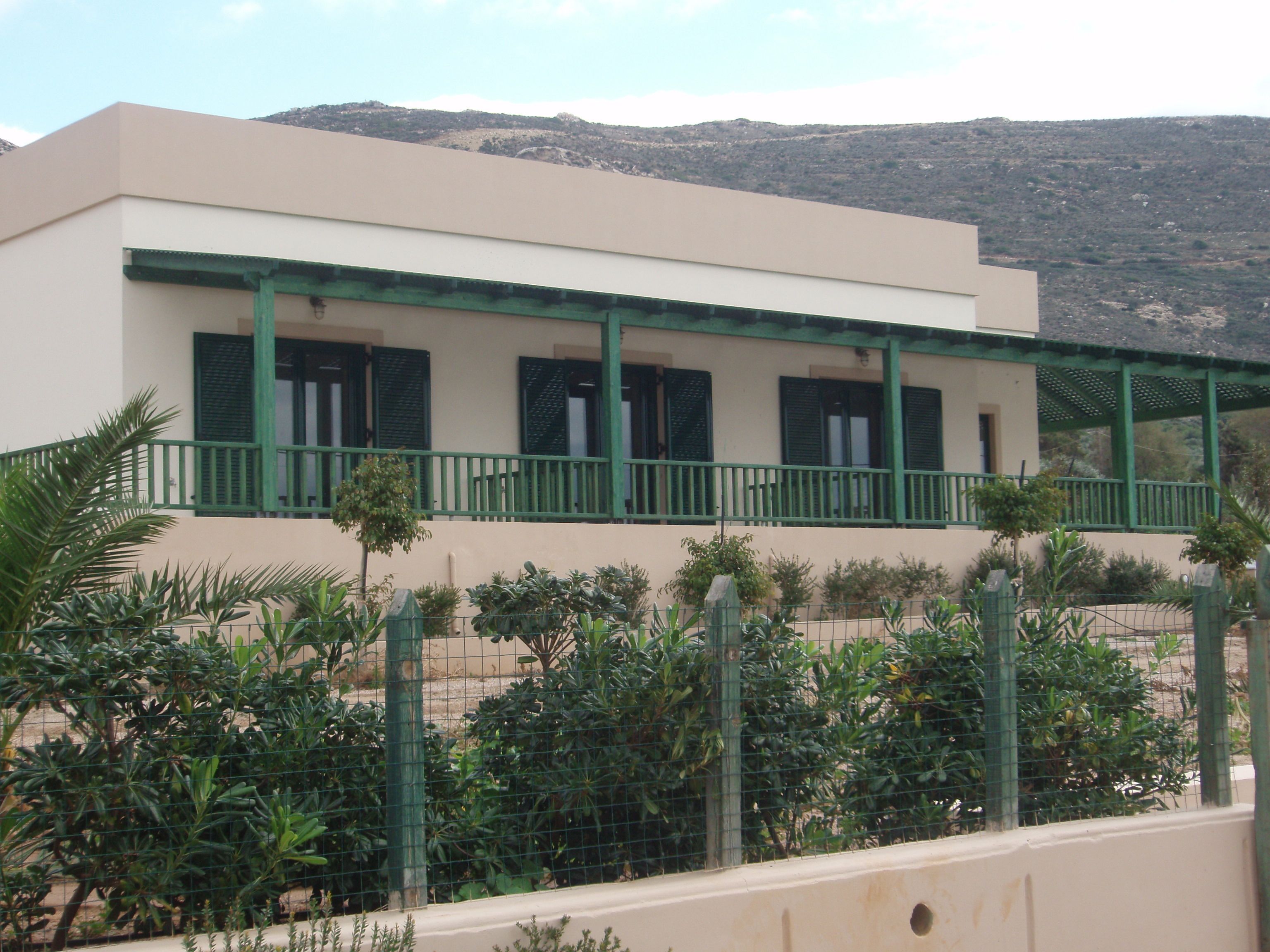 A building with a flat roof, a balcony with green railings, and a mountainous background.