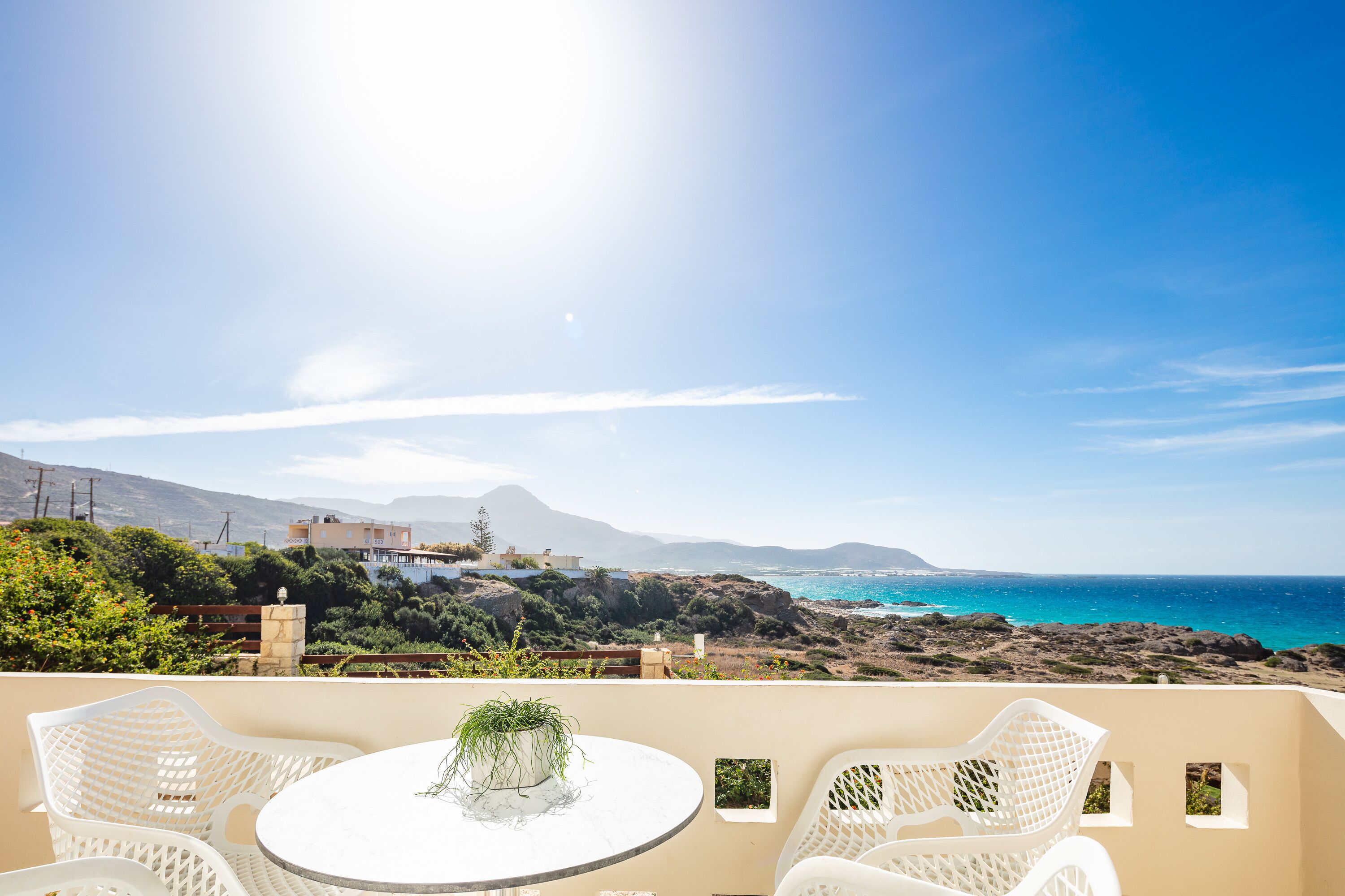 A terrace with a round table and white chairs, overlooking a coastal landscape with mountains and the sea.