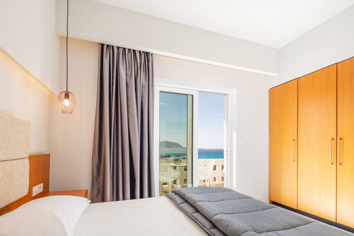 Une chambre d’hôtel moderne, dotée d’un lit, d’une armoire en bois, d’un balcon avec vue sur la mer et d’un luminaire suspendu.