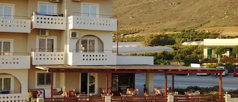 A multi-story building with balconies, a covered outdoor seating area with tables and chairs, and a stone wall in the foreground.