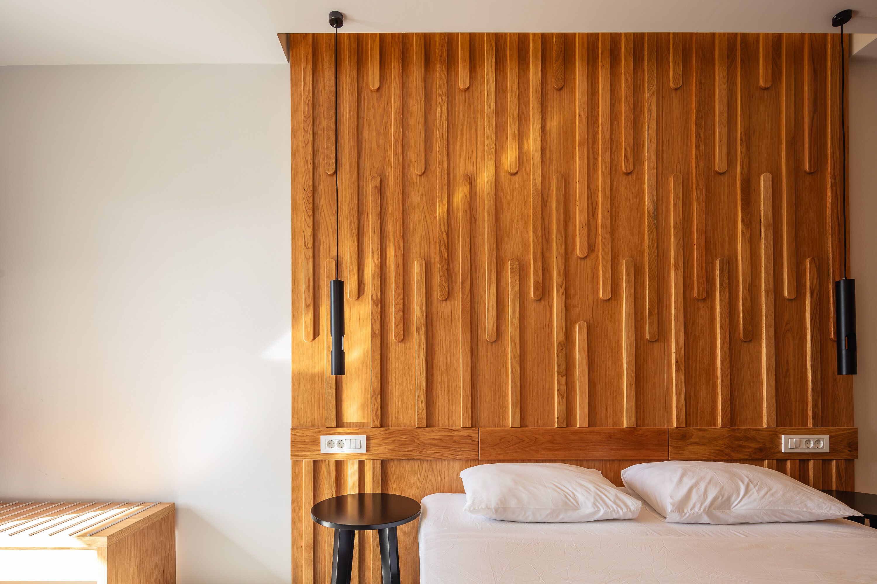 Une chambre à coucher avec une tête de lit en bois, un lit recouvert d’une literie blanche, une petite table ronde et un luminaire fixé au mur.
