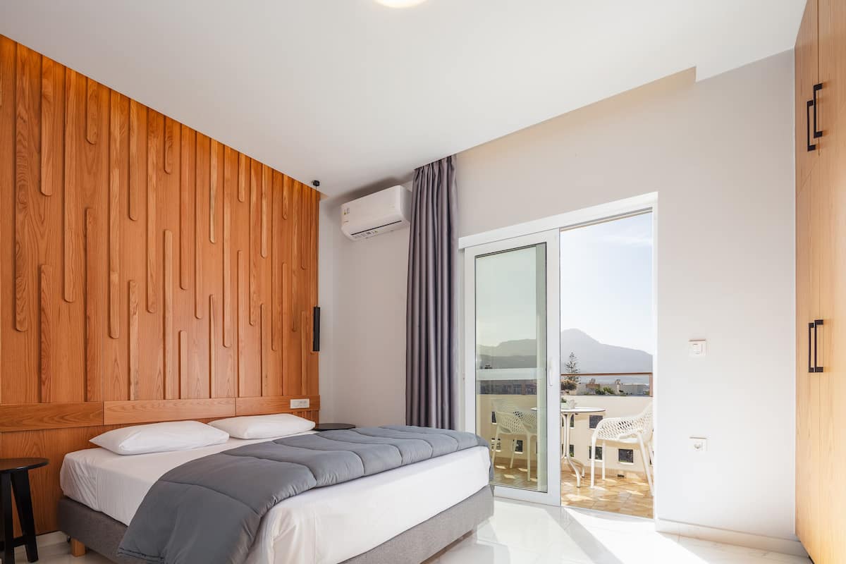 Une chambre avec une tête de lit en bois, un lit avec une literie grise, une porte-fenêtre coulissante donnant sur un balcon et une vue sur l’extérieur.