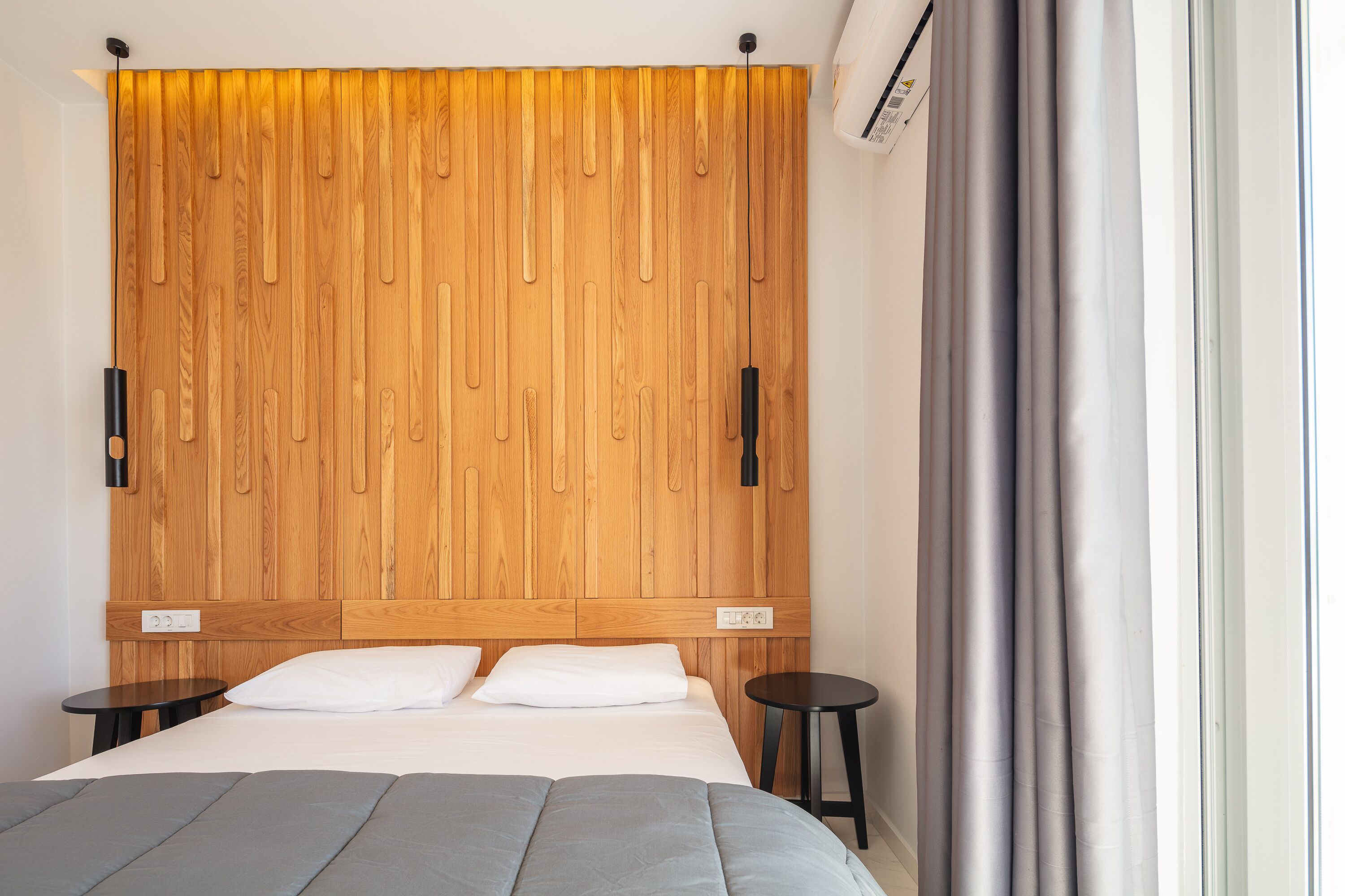 Een slaapkamer met een houten hoofdbord, een bed met grijze beddengoed, twee nachtkastjes en een raam met grijze gordijnen.