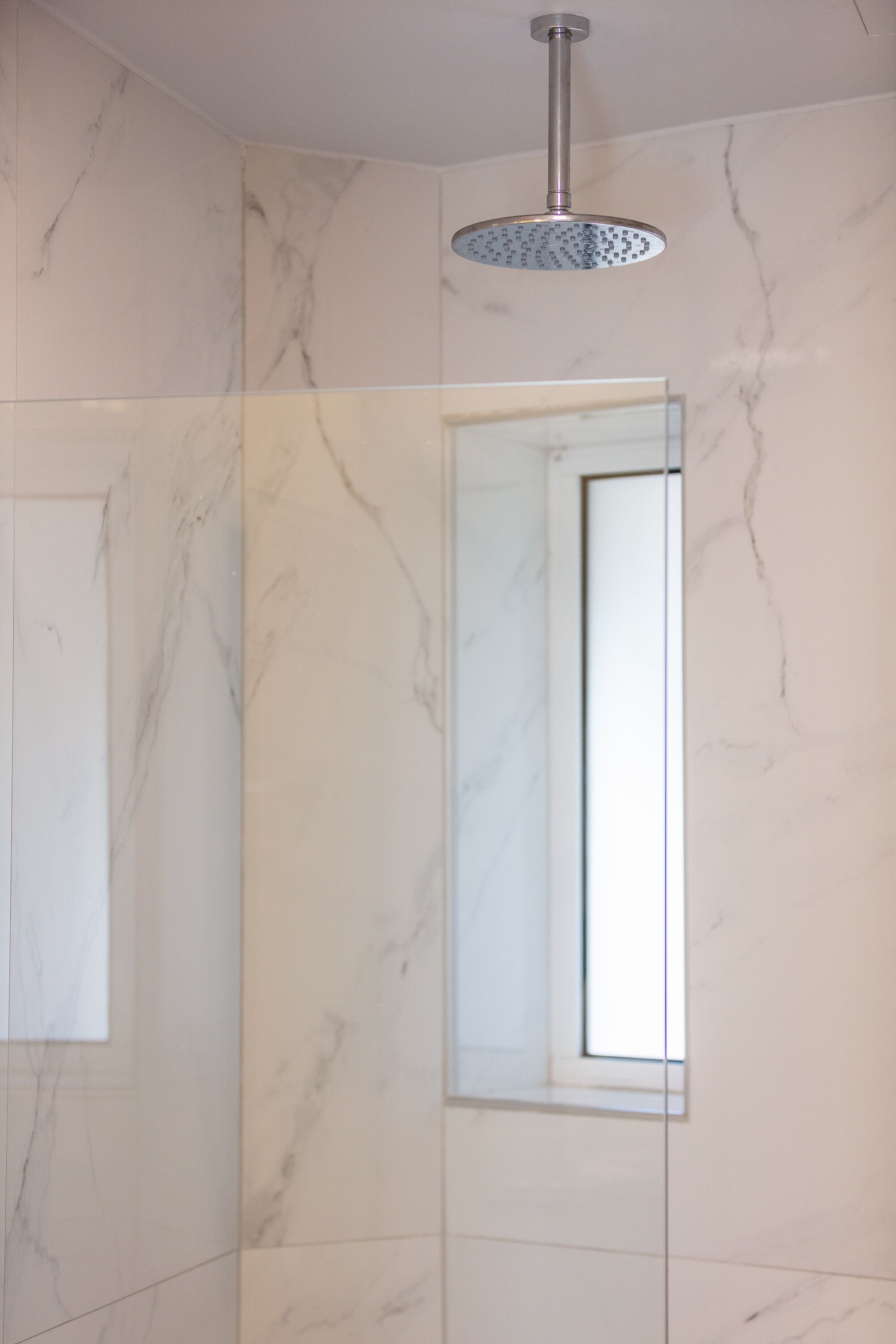 A bathroom with marble walls, a shower with a rain showerhead, and a mirror reflecting part of the room.