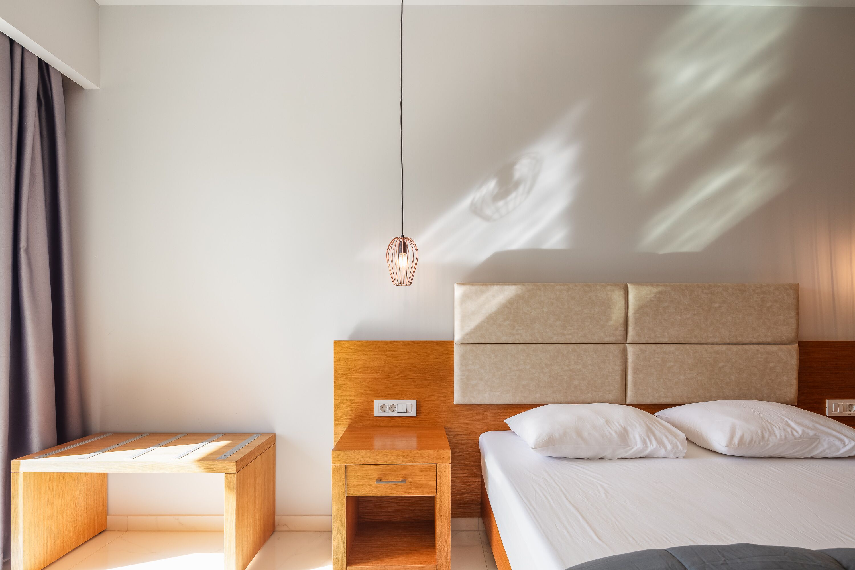 A modern hotel room with a wooden nightstand, a bedside table, a bed with white linens, and a headboard with a light fixture above.