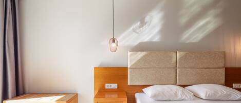 Una habitación de hotel moderna con una mesita de noche de madera, una lámpara de noche y una cama con ropa blanca, con un cabecero que tiene una lámpara colgante.