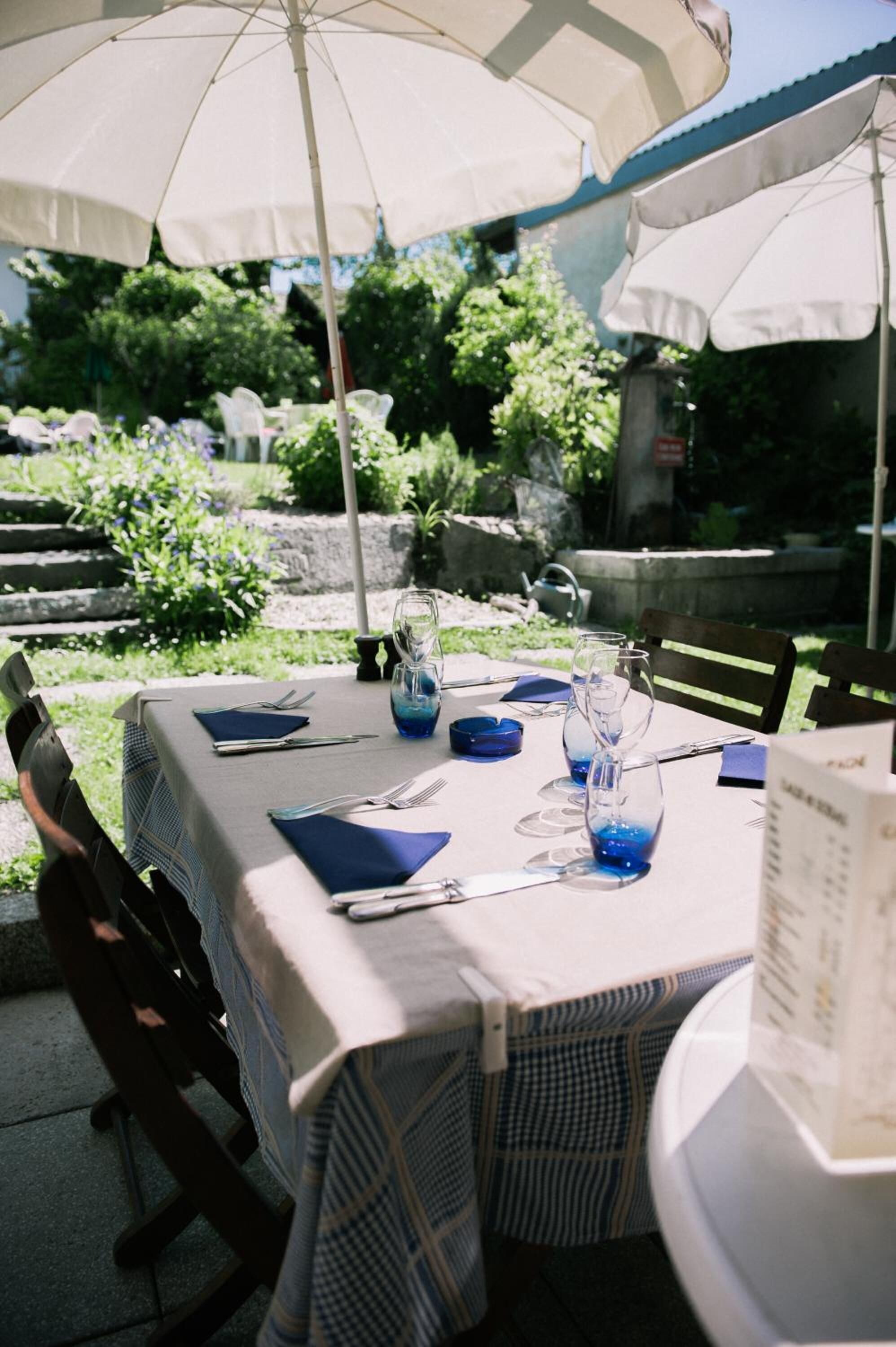An outdoor dining area with tables set for two, covered by white umbrellas, and surrounded by greenery.