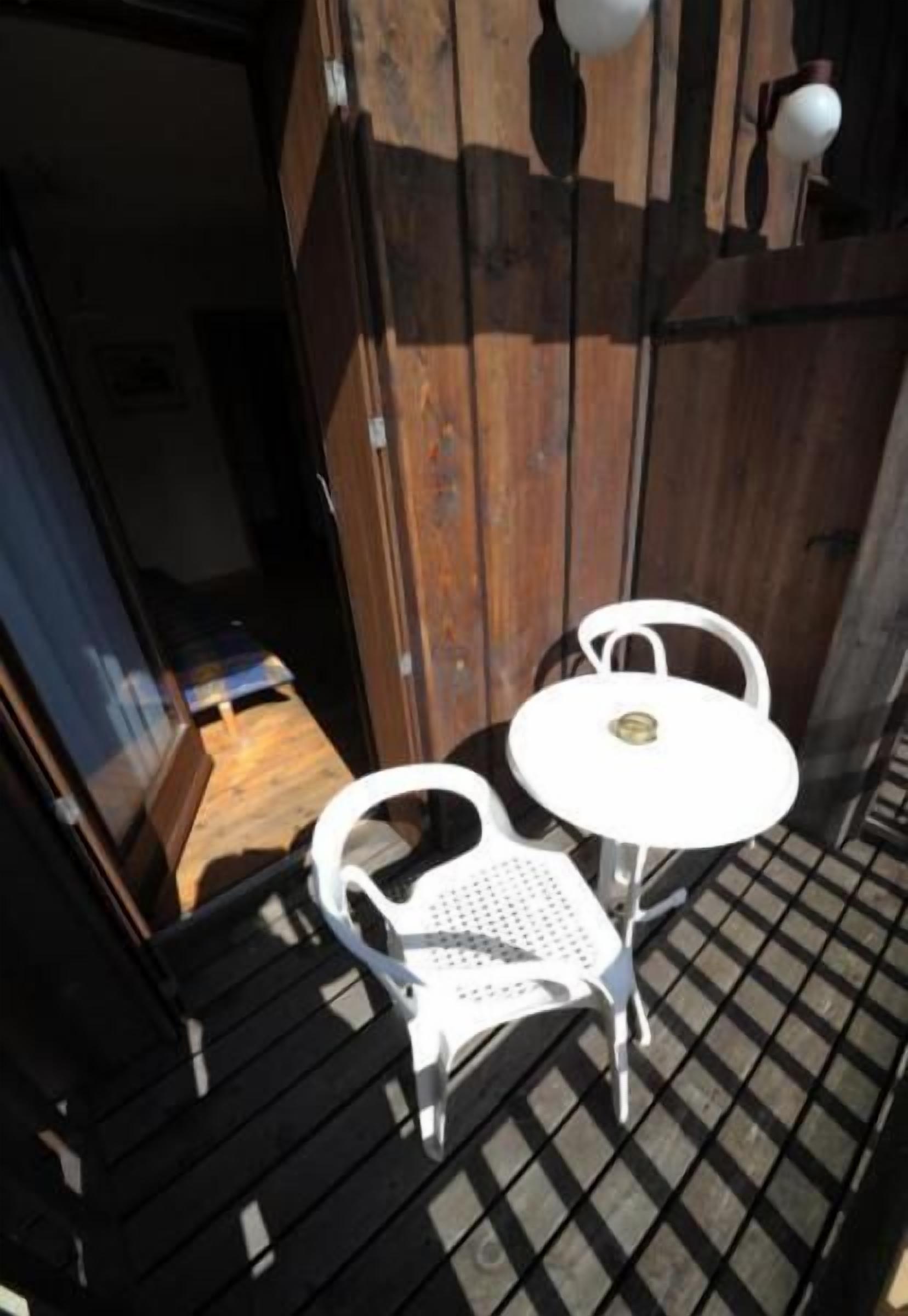 Un petit espace extérieur aménagé avec une table en métal et deux chaises blanches, une clôture en bois en arrière-plan et une porte donnant sur un espace intérieur.
