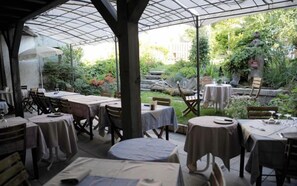 An outdoor patio with tables and chairs, surrounded by greenery and a stone wall.
