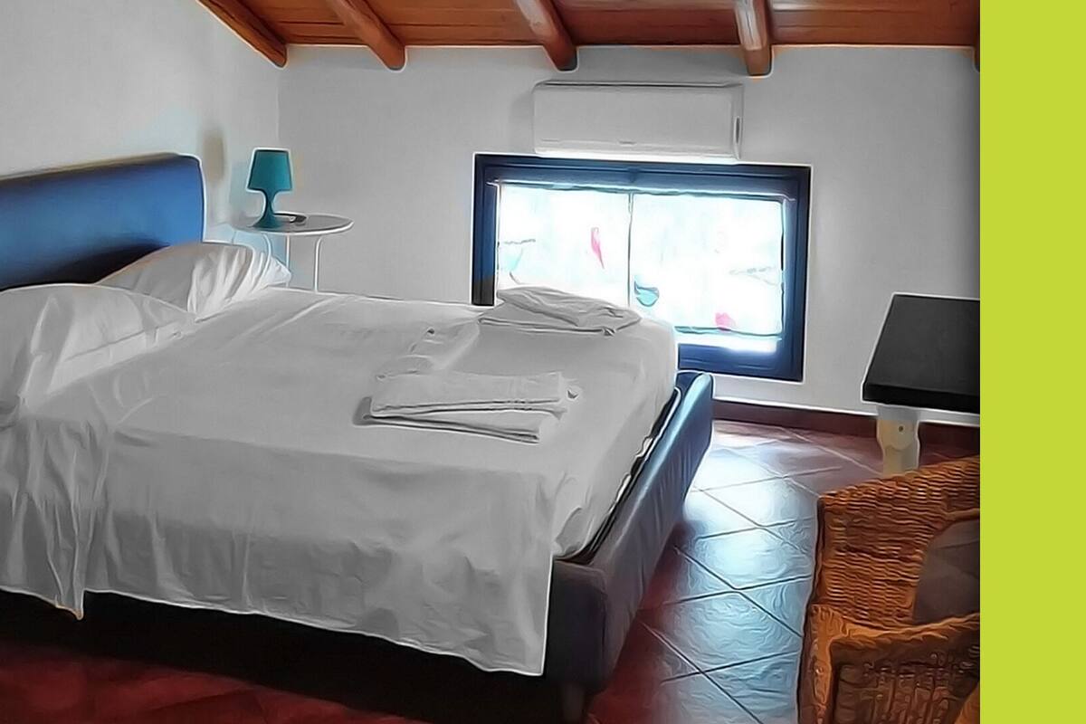 Une chambre à coucher avec un lit, une table de chevet, une lampe, une fenêtre et une chaise en osier.