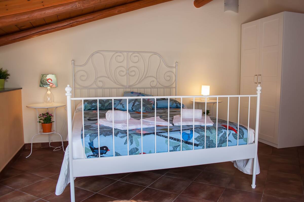 Un lit en métal blanc, doté d’une tête de lit décorative, se trouve dans une pièce dont le sol est recouvert de carreaux et le plafond est en bois.