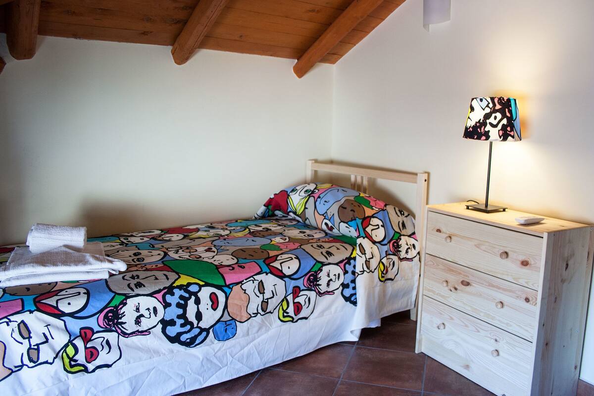 Une chambre à coucher avec un lit recouvert d’une couverture colorée, une table de chevet en bois avec une lampe et un mur blanc.