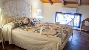 A neatly made bed with a patterned bedspread, a bedside table with a lamp, and a window with a view of chairs outside. - Le Coccole (Catania)