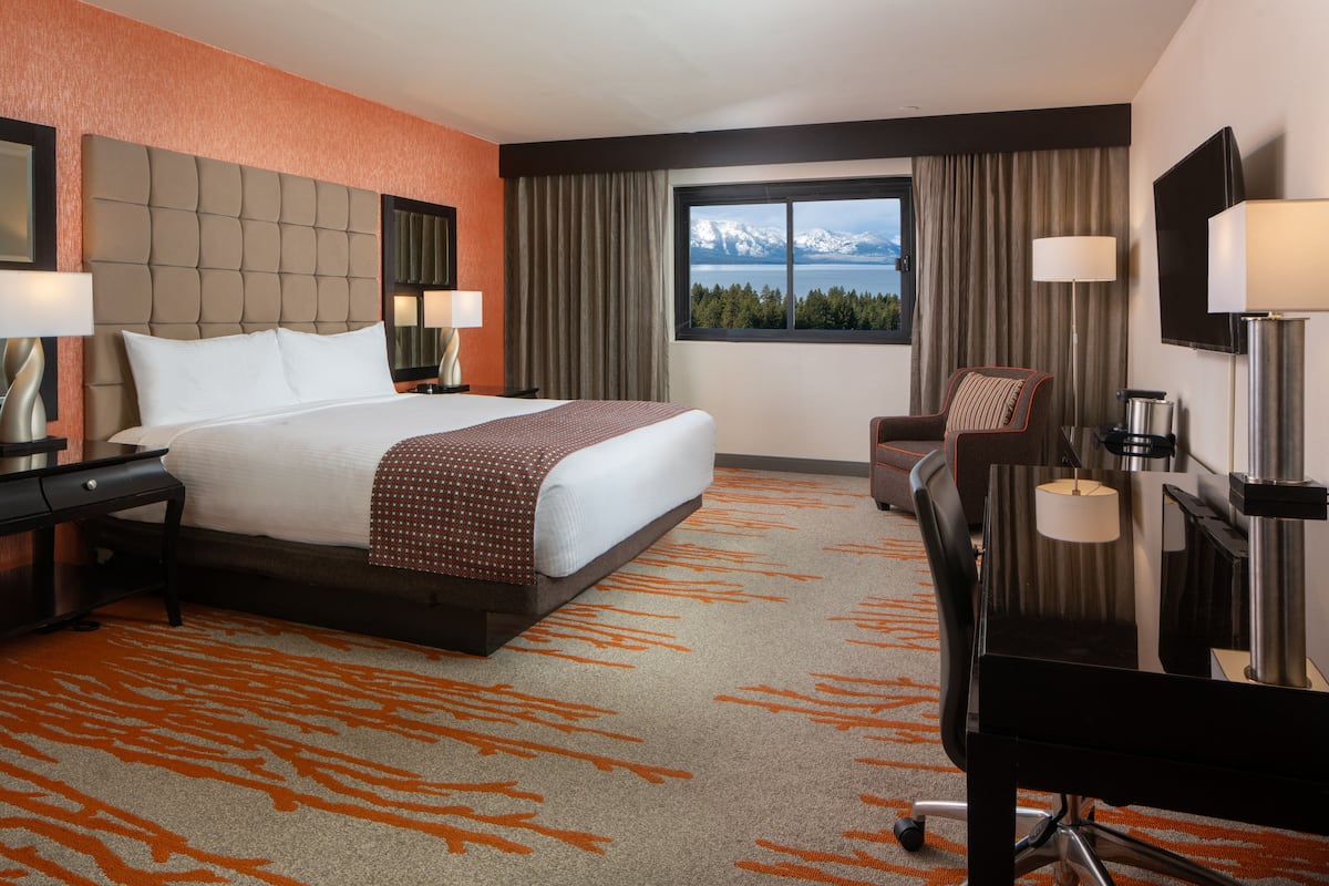 A hotel room with a large bed, a desk, a chair, a TV, and a window with a mountain view.