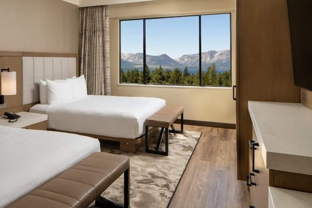 A hotel room with two beds, a desk, a chair, a large window with a mountain view, and a bathroom.