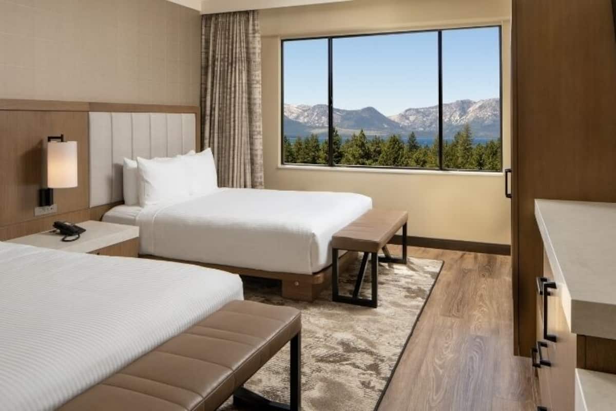 A hotel room with two beds, a desk, a chair, a large window with a mountain view, and a bathroom.