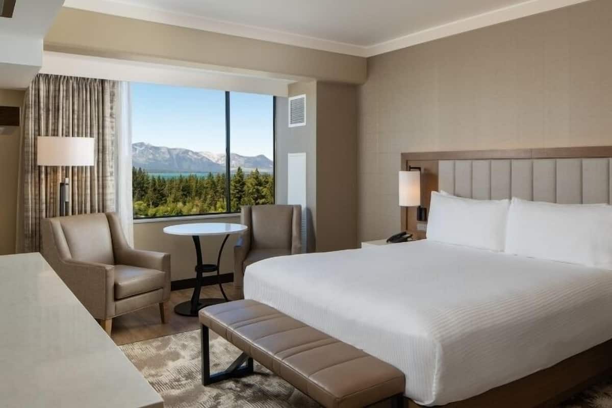A hotel room with a large bed, a desk, a chair, a small table, and a view of mountains.