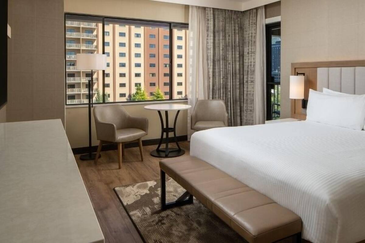 A hotel room with a large bed, a desk, two chairs, a small table, and a bench.