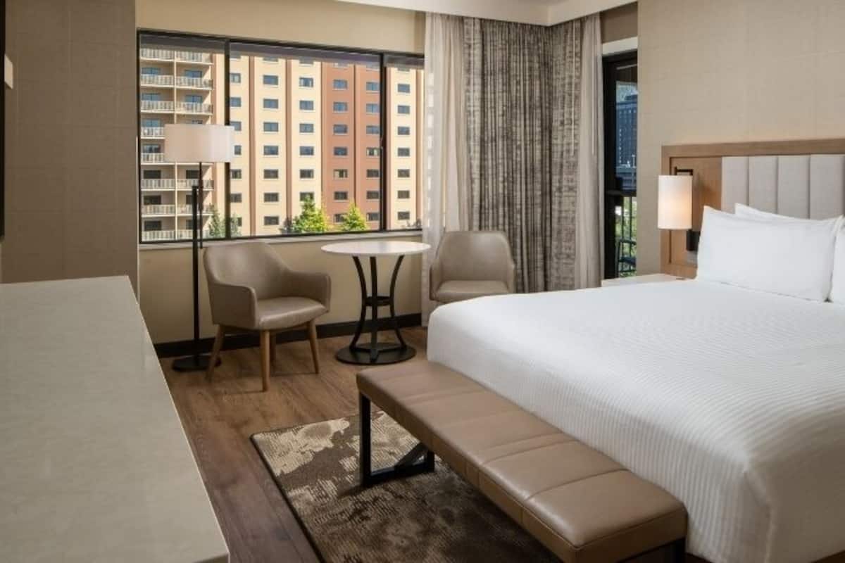 A hotel room with a large bed, a desk, two chairs, a small table, and a bench.
