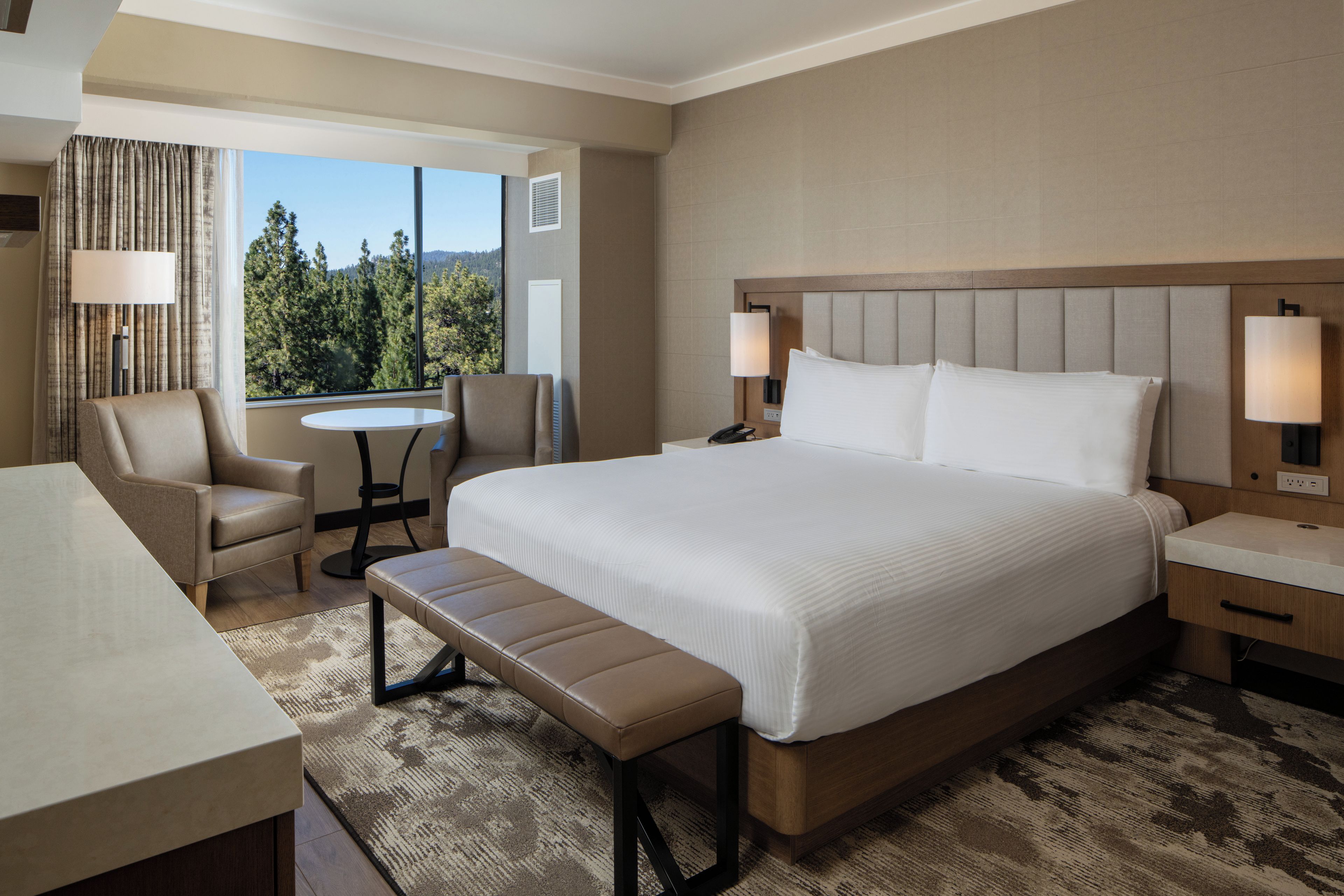 A hotel room with a large bed, a desk, a chair, a small table, and a bench.