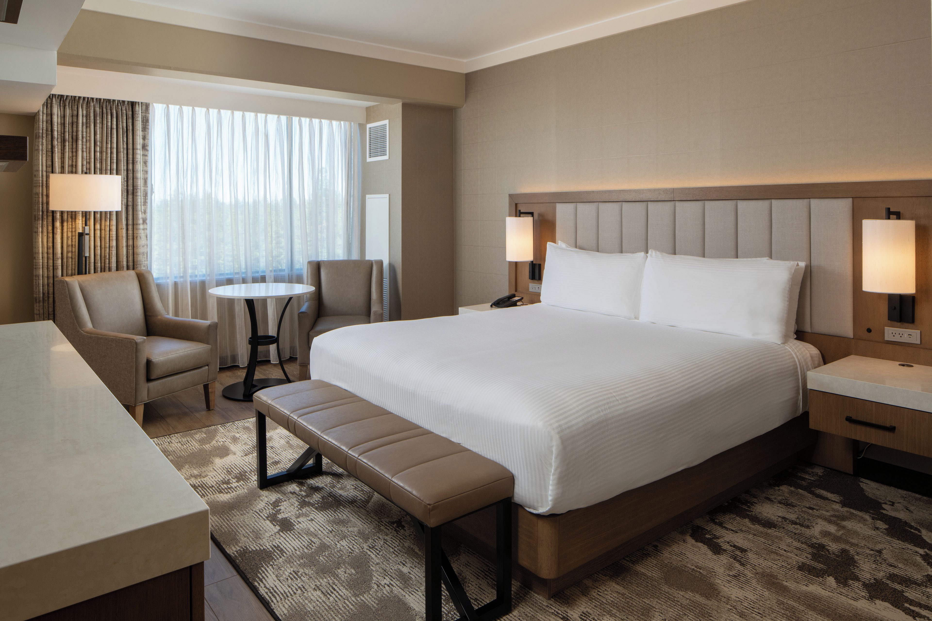 A hotel room with a large bed, a desk, a chair, a small table, and a bench.