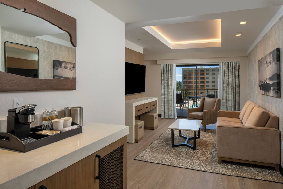A modern hotel room with a kitchenette, a flat-screen TV, a sofa, and a balcony with a city view.