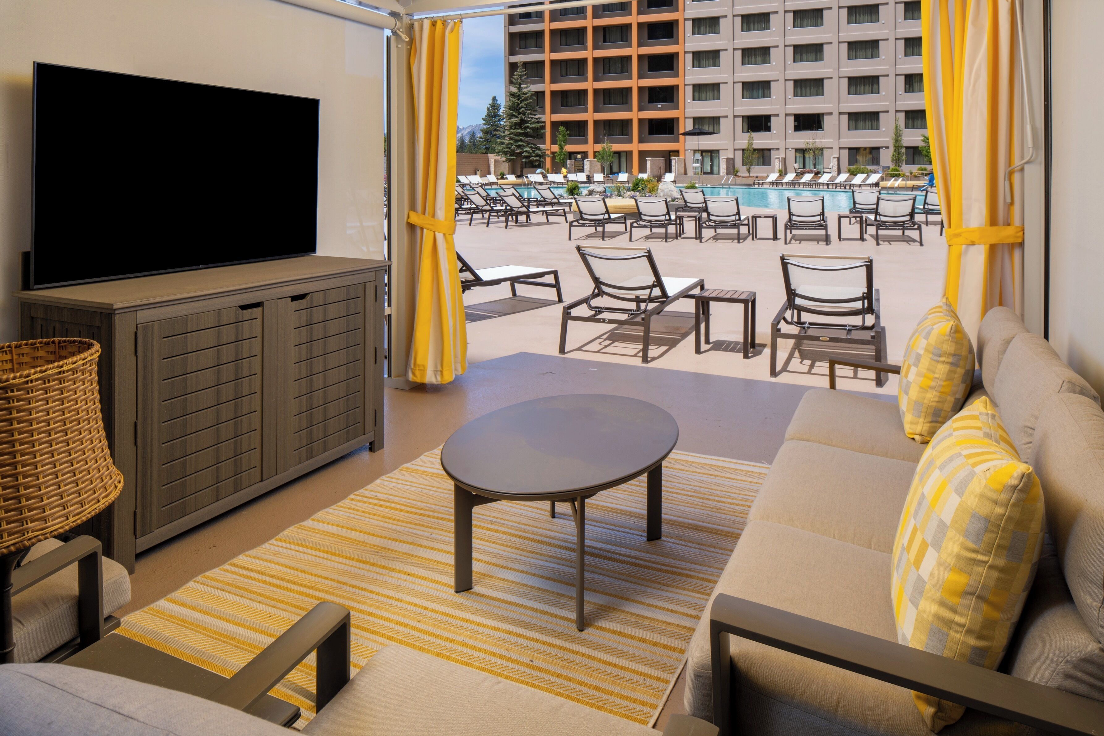 A living room with a flat-screen TV, sofa, and armchair, a small table, and a view of a pool and hotel building.