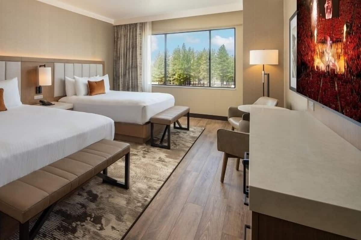 A hotel room with two beds, a desk, a chair, and a large window with a view of trees.