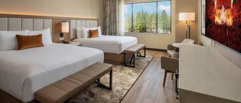 A hotel room with two beds, a desk, a chair, and a large window with a view of trees.
