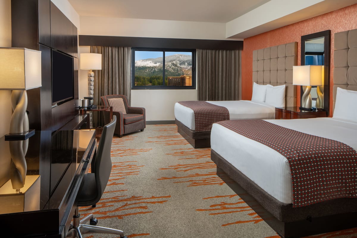 A hotel room with a large bed, a smaller bed, a desk with a TV, and a view of mountains through the window.
