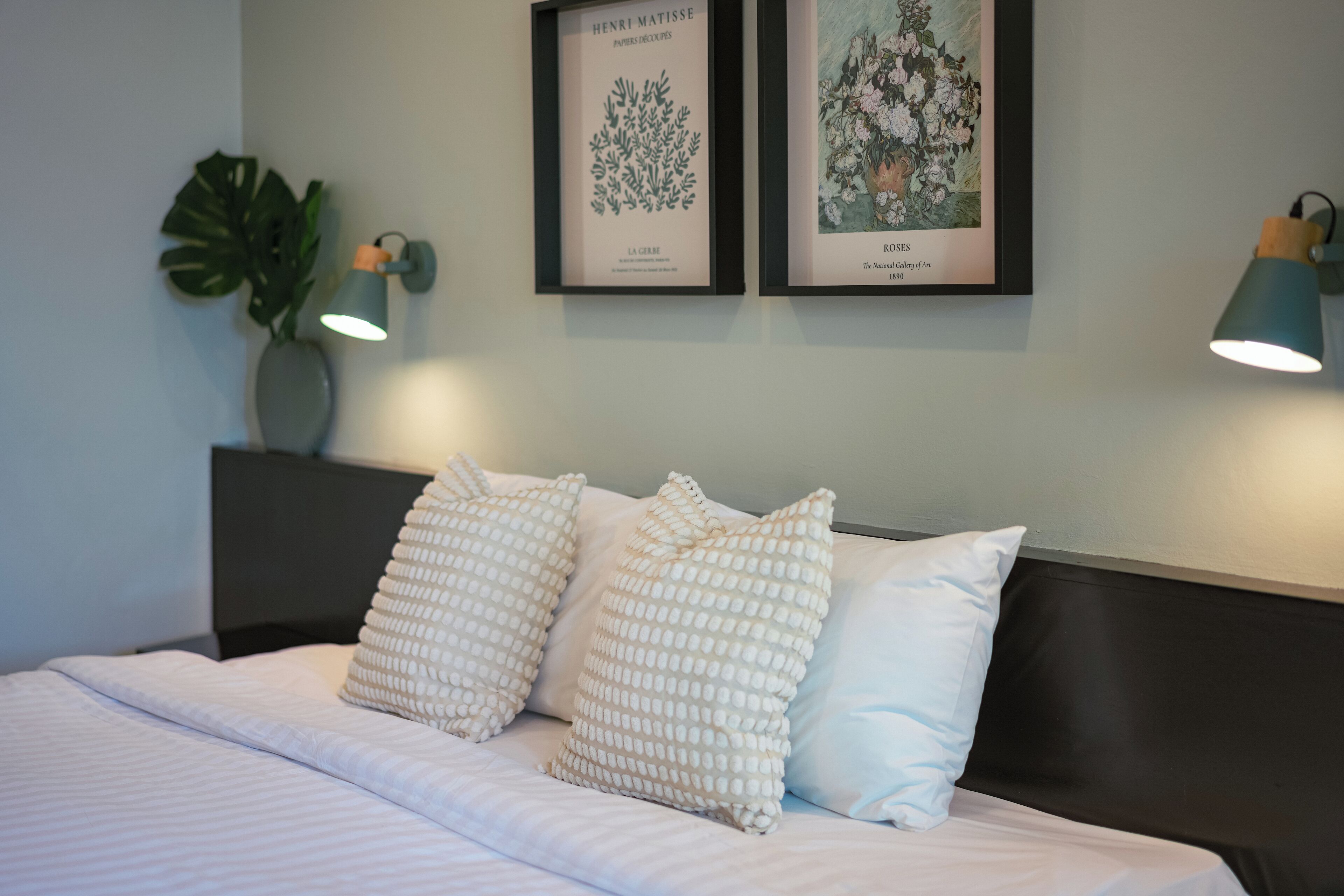 A neatly made bed with white bedding and patterned pillows, a dark headboard, and two framed artworks above.