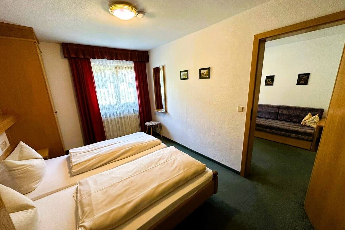 Ein Hotelzimmer mit zwei Betten, einem Fenster mit Vorhängen, einem kleinen Hocker, einem Spiegel und einem Sofa im angrenzenden Raum.