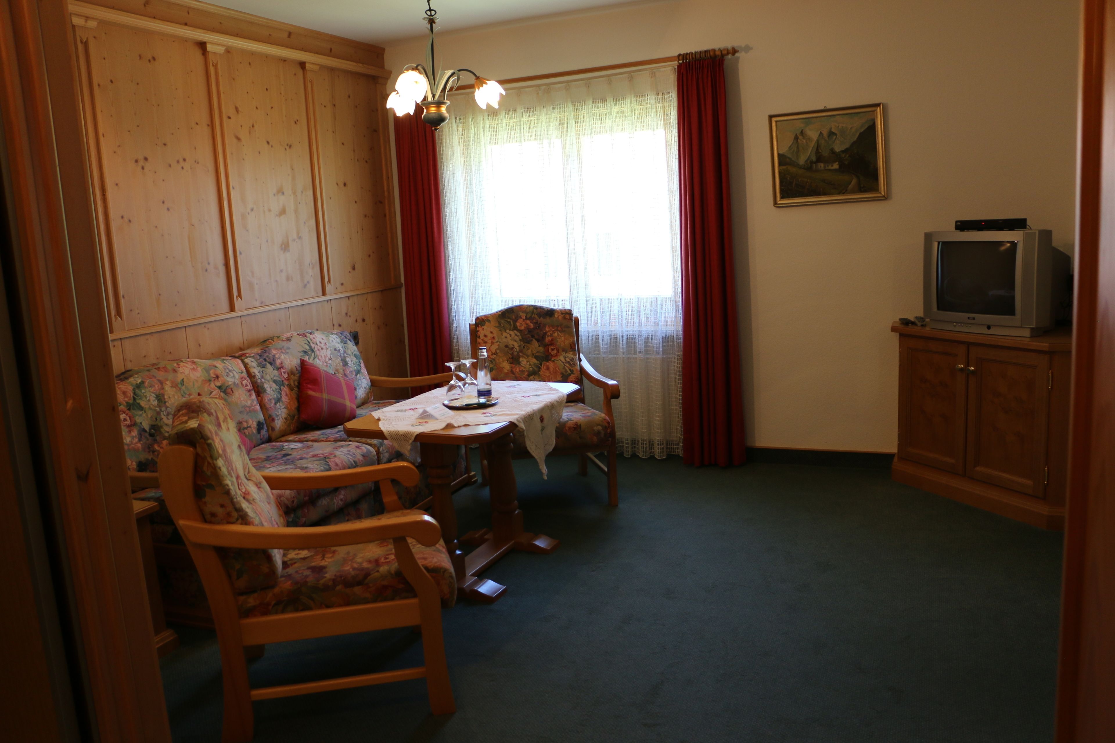 A living room with a wooden sofa, floral armchairs, a small table, a television on a cabinet, and a painting on the wall.