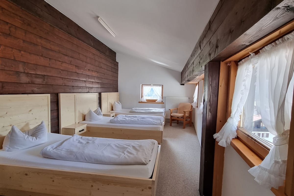 Ein Zimmer mit drei Holzbetten, einer Holzzahlendecke, einem Fenster mit weißen Vorhängen und Blick auf ein Gebäude.