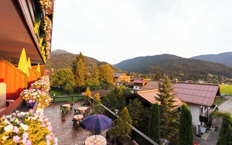 A terrace with outdoor seating, surrounded by flowers and trees, overlooking a mountainous landscape.