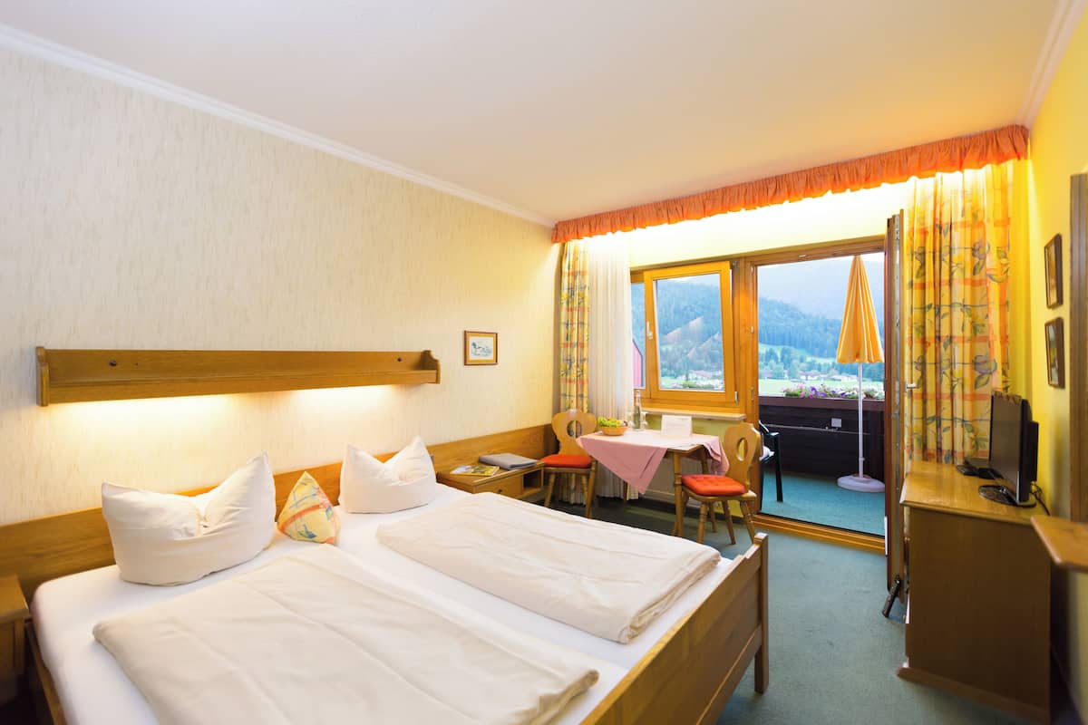Ein Hotelzimmer mit einem Doppelbett, einem Schreibtisch mit Computer, einem Stuhl, einem kleinen Tisch mit Pflanze und Blick ins Freie durch ein Fenster.