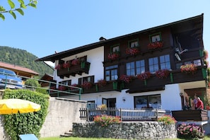 Un edificio de dos pisos con balcones decorados con flores rojas, un muro de piedra en el frente y una valla de madera.