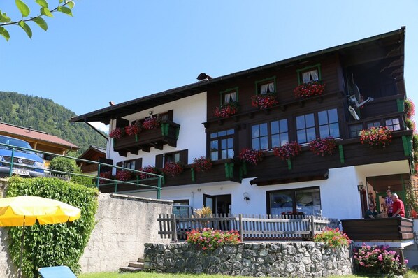 Un edificio de dos pisos con balcones decorados con flores rojas, un muro de piedra en el frente y una valla de madera.