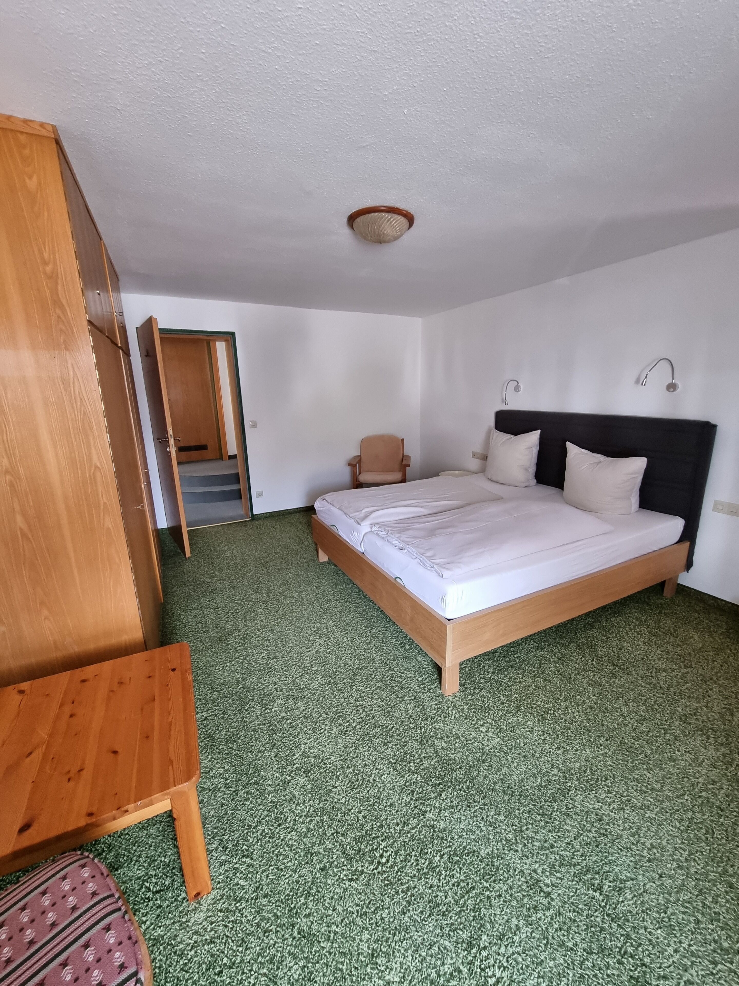 A bedroom with a wooden bed, a chair, a wardrobe, and a door leading to a staircase.