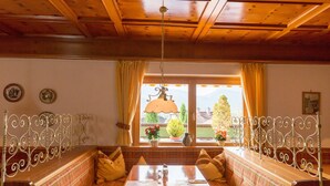 A cozy room with a wooden ceiling, a bench with cushions, a table with a metal railing, and a window with curtains. -  Hotel Sonnleiten (Reit im Winkl)