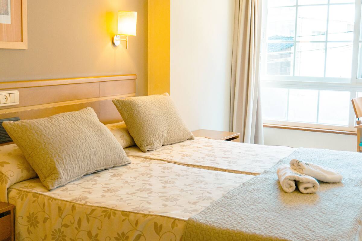 A hotel room with two beds, a nightstand, a lamp, and a window with curtains.