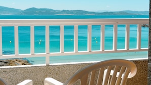 Ein Balkon mit weißer Brüstung, der auf einen Strand mit klarem, blauem Wasser und ferne Hügel blickt.