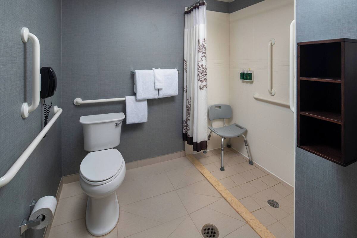 Une salle de bain équipée de toilettes, de barres d’appui, d’une douche avec rideau, d’une barre de maintien et d’une petite chaise.