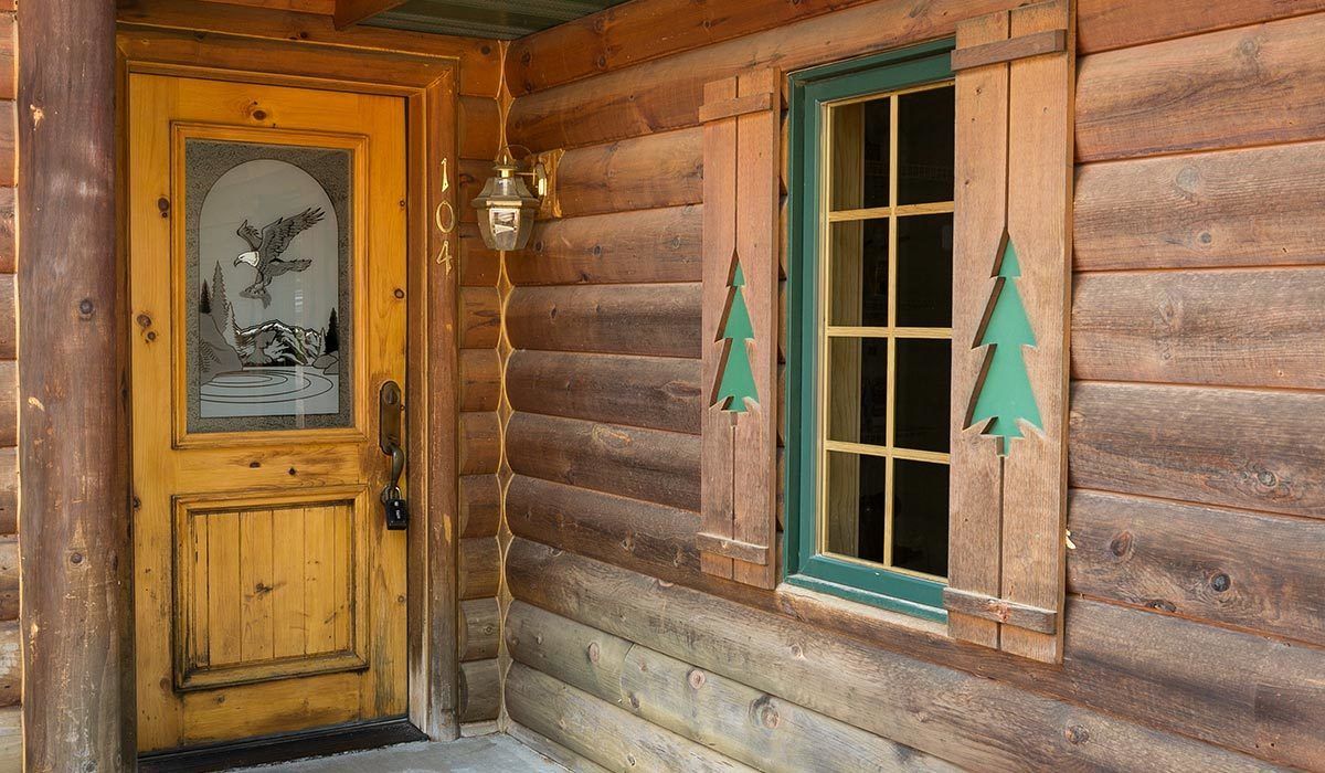 A wooden cabin with a glass door featuring a mountain scene, a green-framed window, and a wall-mounted light fixture.