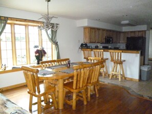 A dining area with a wooden table and chairs, a kitchen area with wooden cabinets, and a view through a window with curtains. - Log Gables - Lake Front (Wisconsin Dells)