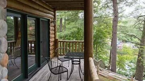 Un terrazzo in legno con tavolo e sedie, un muro in pietra e vista su alberi e un edificio sullo sfondo.