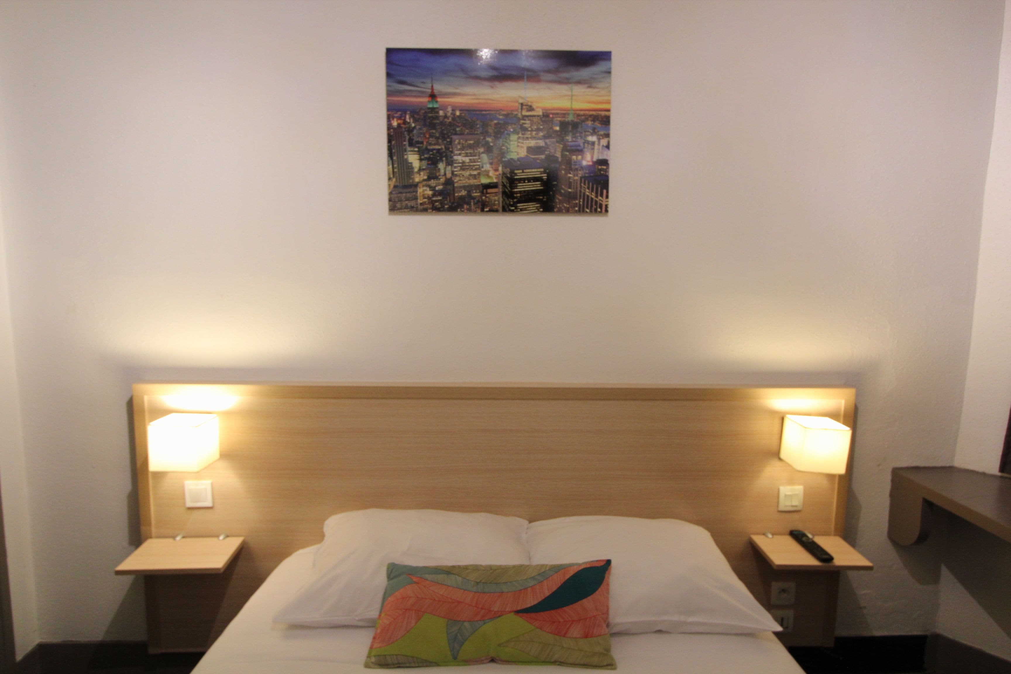 A hotel room with a bed, two bedside lamps, a nightstand, and a cityscape painting on the wall.