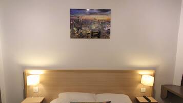 A hotel room with a bed, two bedside lamps, a nightstand, and a cityscape painting on the wall.