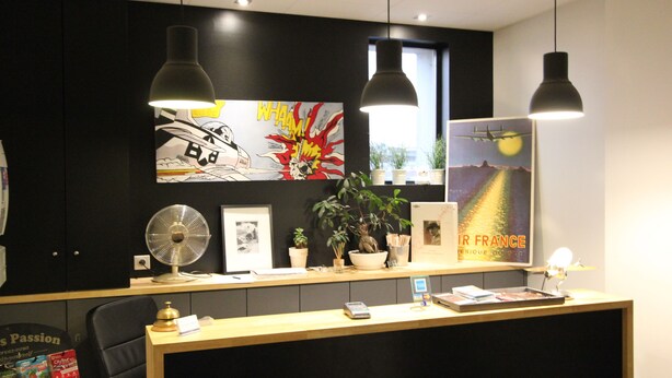 A reception desk with a black and white comic-style artwork, a poster of a landscape, and a fan. - Hotel Vol de Nuit (Toulouse)