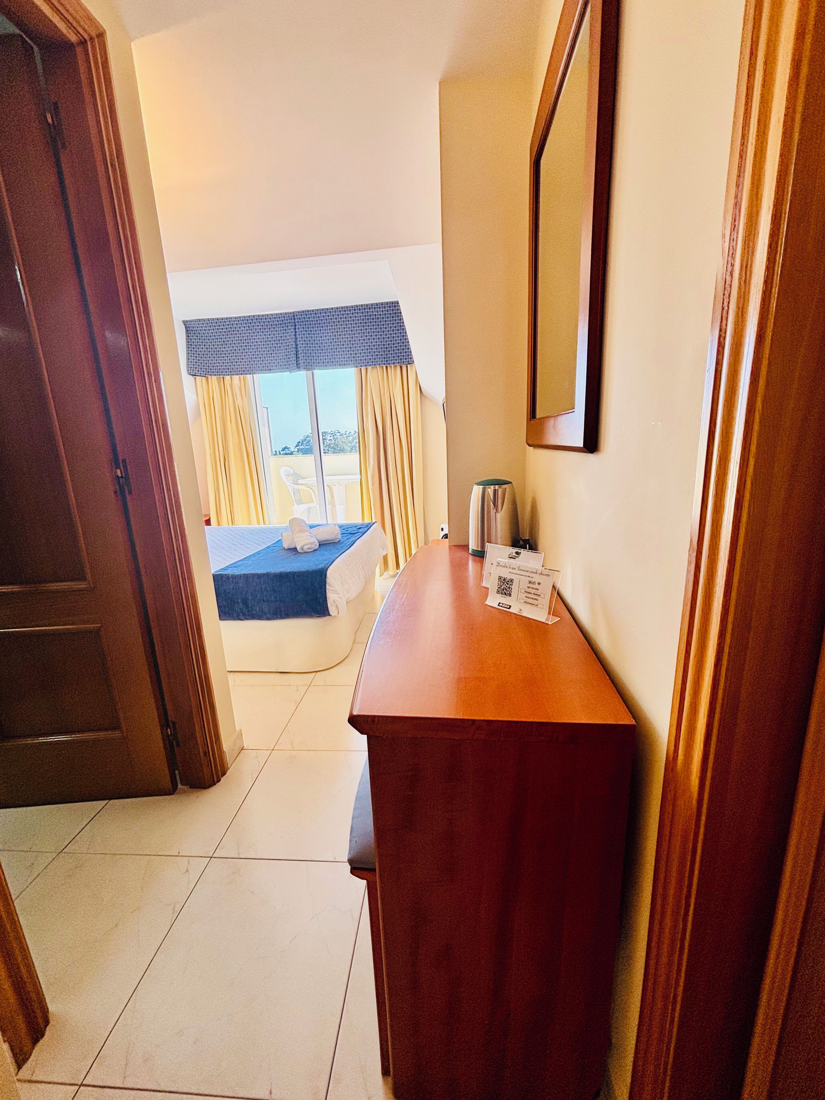 A hotel room with a wooden desk, a bed with a blue headboard, a window with curtains, and a view of the outside.